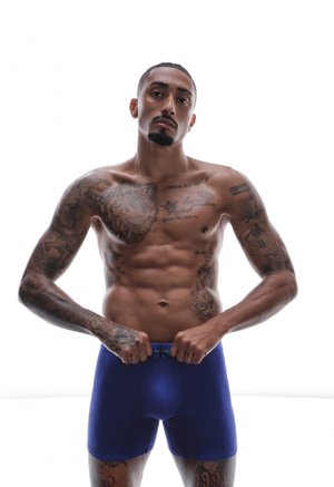 Krekls bez krekla, vīrietis ar vairākām tetovējumiem pielāgo zilas bokseršortus baltā fonā, skatoties tieši uz priekšu ar neitrālu izteiksmi.