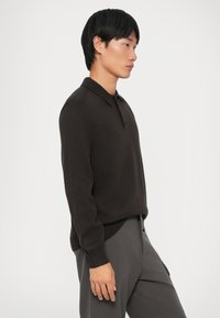 Suéter de polo marrón de punto con cuello, que presenta un acabado texturizado y un corte relajado, combinado con pantalones grises.