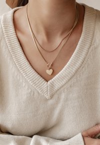 Gelaagde gouden halskettingen met een hartvormige hanger, tegen een textuur van een crèmekleurige trui met een V-hals ontwerp.