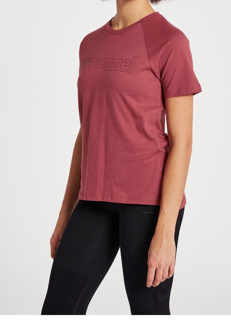 Femme portant un T-shirt de sport Hummel bordeaux à manches courtes et un legging noir, posant avec les bras détendus le long du corps.