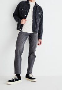 Donkergrijze spijkerjas, knoopjes met borstzakken. Witte T-shirt eronder, gecombineerd met grijze jeans en zwart suède sneakers met witte zolen.