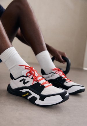 Person iført hvide New Balance Minimus sneakers med røde snørebånd og hvide sokker, der holder en kettlebell, mens de sidder på gulvet.