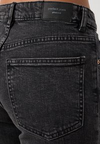 Black denim jeans with a classic cut, featuring a back pocket and branded leather tag. The texture appears slightly distressed.