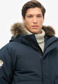 Marineblå isoleret parka med hætte med pelskant, lynlås foran og en ribstrikket sweater under. Udstyret med mærkat på ærmet.
