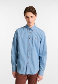 Lichtblauw, langemouwshirt van denim; heeft een knoopsluiting bij de kraag, bruine knopen en een klein geborduurd logo op de borst.