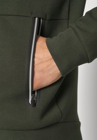 Grön sweatshirt med en dragkedjeficka med svarta detaljer och silveraccent. Materialet verkar vara strukturerat och mjukt.