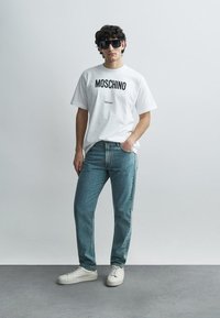 T-shirt branca de algodão com "MOSCHINO Milano" impresso a preto, combinada com jeans de ganga azul clara e sapatilhas brancas. Óculos de sol pretos completam o visual.