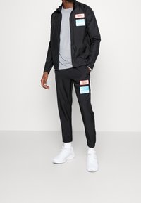 Man in een zwarte Nike trainingspak met rode en blauwe patches, een grijs t-shirt, witte sokken en witte sneakers, staand tegen een witte achtergrond.