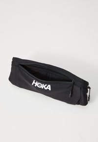 Musta vetoketjullinen pussi, joka on valmistettu teksturoidusta kankaasta ja jossa on valkoinen logo. Kompakti, suorakulmainen muoto, joka sopii säilytettäväksi.
