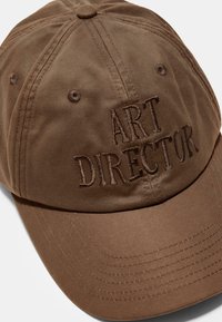 Brun bomuldsbaseballcap med broderet tekst "ART DIRECTOR." Har seks-panel design og syede detaljer på skyggen.