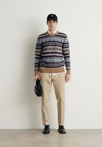 Strickpullover mit mehrfarbigen geometrischen Mustern, beige gerippter Saum, beiger Hose und schwarzen Schuhen. Schwarze Kappe und kleine Schultertasche.