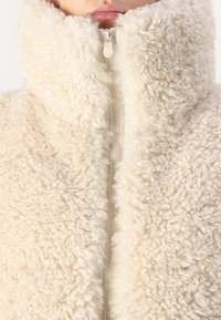 Borg-Woll-Zip-Jacke in weichem Creme. Verfügt über einen hohen Kragen und eine strukturierte, flauschige Oberfläche mit einem elfenbeinfarbenen Reißverschlussdetail an der Vorderseite.