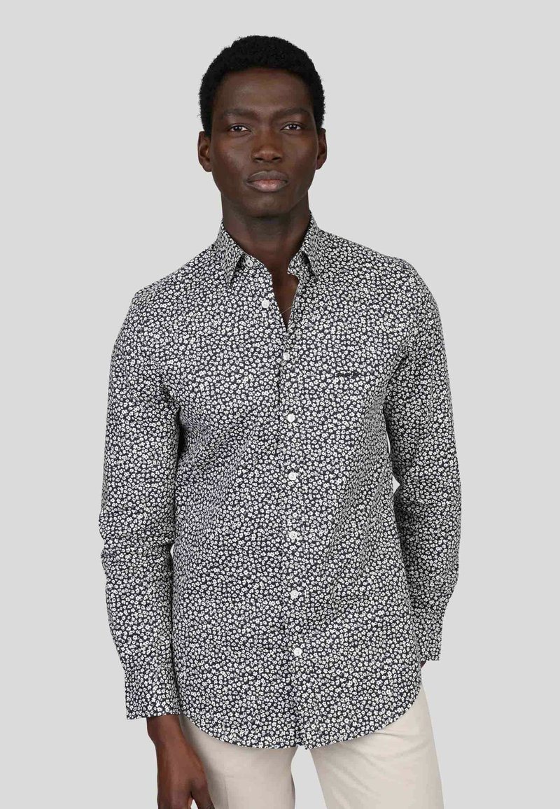 Chemise à manches longues noire avec imprimé floral blanc, col boutonné et poche poitrine. Le tissu semble léger avec une texture lisse.