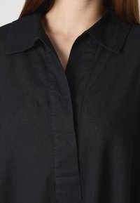 Camisa de linho preta com colarinho, apresentando uma abertura com pala e uma superfície ligeiramente texturizada. A vista em close realça os detalhes do tecido.