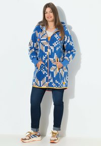 Cappotto in pile con cappuccio di colore blu con motivi geometrici, dettagli arancioni, chiusura a zip e tasche laterali. Abbinato a jeans scuri e sneakers multicolori.