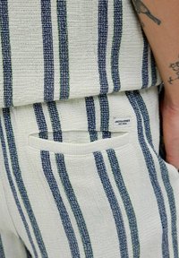 Könnyű, krémszínű ruhadarab, tengerészkék és zöld függőleges csíkokkal. Jellemzője egy kis címke és egy oldalzseb a nadrágon.