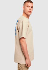 Beige bomulds t-shirt med løs pasform, korte ærmer og fed blå grafisk tekst på højre side. Glat tekstur, minimalistisk design.