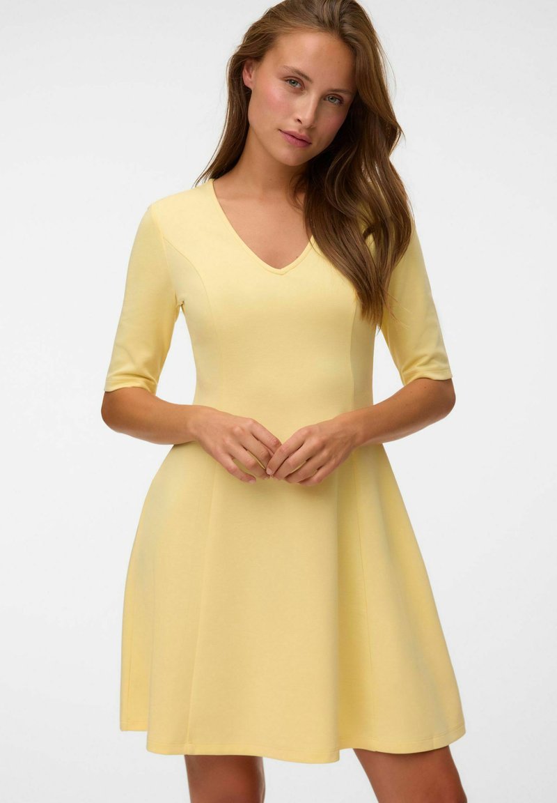 Femme aux longs cheveux bruns portant une robe jaune au genou avec des manches mi-longues et un col en V, debout les mains doucement jointes.