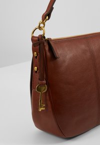 Bruine leren handtas met een gladde textuur, voorzien van een ritsluiting, gouden hardware en een sleuteldetail dat aan de band is bevestigd.