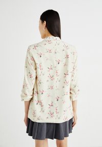 Blazer à imprimé floral en crème avec des fleurs rose pâle et vertes, manches longues retroussées jusqu'aux coudes, design col, tissu doux, détail de couture au dos.