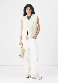 Kremfarget quilted vest med en lapp lomme, båret over en mørk topp. Kombinert med vide hvite bukser og lyse sneakers. Har en liten veske.