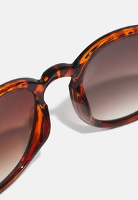 Schildpatt-Sonnenbrille mit einem dicken, runden Rahmen und braunen Verlaufsgläsern. Die Oberfläche ist glatt mit einem glänzenden Finish.