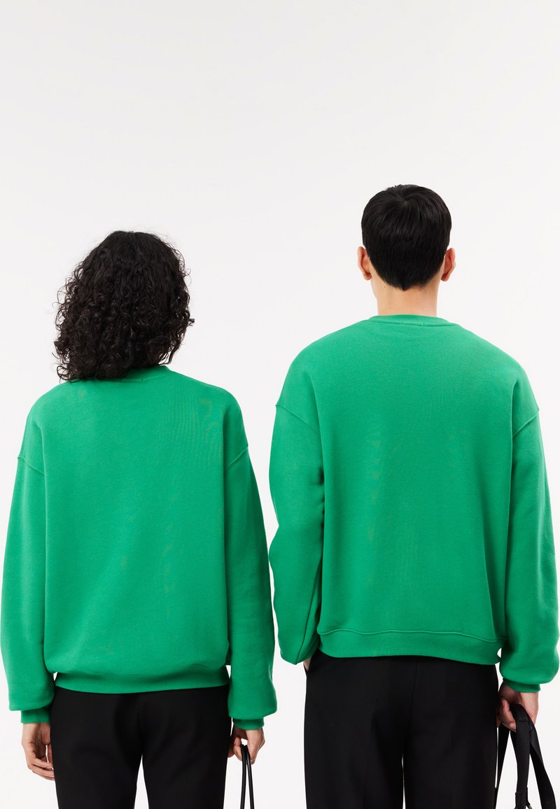 Sweatshirts verts à col rond avec des épaules tombantes, des poignets et des ourlets côtelés, portés par deux personnes avec des pantalons noirs et des accessoires.