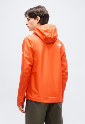 Ung person iført lys oransje hettejakke med ryggen mot, stående mot enkel lys bakgrunn.