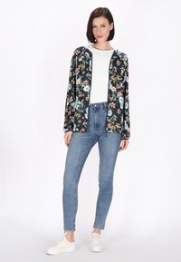 Floral zwart ritsjasje met lange mouwen, gedragen over een witte top, gecombineerd met blauwe jeans en witte sneakers.