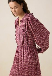 Robe florale violette avec des manches volantes, un col plissé, une taille cintrée et une texture légère, présentant un motif de feuilles répétées.