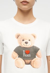 Άνθρωπος που φοράει λευκό πουκάμισο με γραφικό ενός αρκουδάκι που φοράει σκοτεινό πουκάμισο με την επιγραφή «I [κόκκινη καρδιά] Παρίσι».