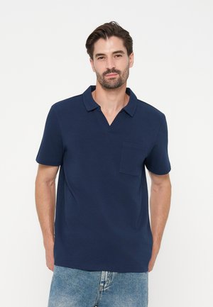 Homme aux cheveux foncés et à la barbe, portant un polo bleu marine à manches courtes et un jean bleu clair, debout devant un fond blanc uni.