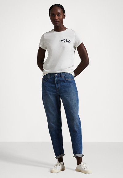 Λευκό βαμβακερό t-shirt με λογότυπο "POLO", σε συνδυασμό με τζιν παντελόνι μέτριας απόχρωσης. Το μοντέλο φορά λευκά αθλητικά παπούτσια, ενώ το τζιν είναι διπλωμένο στα μπατζάκια.