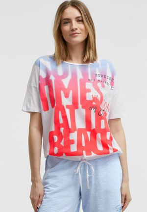 Frau mit schulterlangem blondem Haar, die ein weißes übergroßes T-Shirt mit rotem und blauem Text und hellblaue Shorts mit Kordelzug trägt.