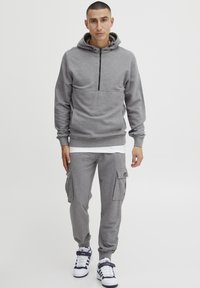 Grijze zip-up hoodie met een voorzak, gecombineerd met grijze cargobroeken met grote zijzakken. Witte t-shirt eronder en witte sneakers.