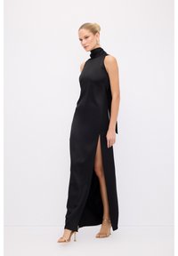 Sort satin maxi kjole med høj hals, ærmeløst design og høj slids til låret. Sættes sammen med gyldne hæle sandaler og minimalistiske øreringe.