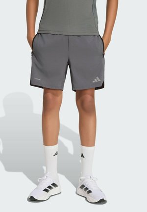 Osoba nosící šedé sportovní šortky s bočními kapsami, bílé ponožky Adidas a bílé tenisky s černými pruhy, stojí s rukama v kapsách.