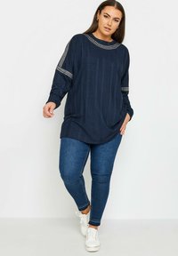 Mörkblå långärmad tunika med strukturerade vertikala ränder och subtila pärldetaljer på axlarna, i kombination med slim-fit jeans och vita sneakers.