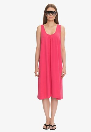 Kobieta w oversize czarnych okularach przeciwsłonecznych, bezrękawowej jasnoróżowej sukience do kolan i czarnych klapkach, stojąca na białym tle.