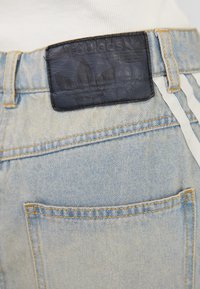 Jean en denim avec un délavage clair, présentant une patch en cuir noir avec logo embossé au dos de la taille et des bandes blanches sur les côtés.