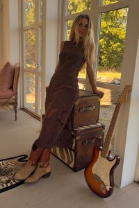 Abito lungo in pizzo marrone con un motivo floreale, abbinato a stivali da cowboy beige. Due bauli vintage marroni e una chitarra elettrica sono nelle vicinanze.