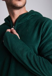 Sweatshirt de felpa verde com um design de meia abertura. Apresenta uma textura suave, mangas longas e um logótipo discreto no peito. O puxador do zíper é metálico.