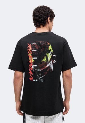 Mand iført sort t-shirt med et farverigt motiv af et nedadvendt hoved og teksten "Dennis Rodman" på ryggen.