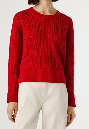 Kobieta ubrana w jasnoczerwony sweter z grubym splotem i wzorzystymi otworami oraz jasnobeżowe spodnie, przedstawiona od szyi do bioder.