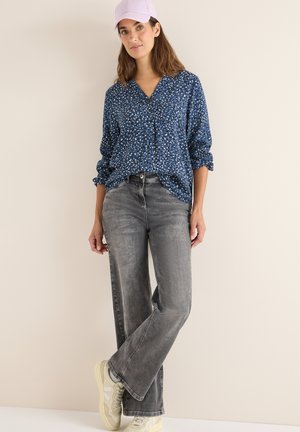 Camicia blu a fantasia con scollatura a V e maniche arrotolate, abbinata a jeans grigi a gamba larga e sneakers chiare. Cappellino lavanda in evidenza.