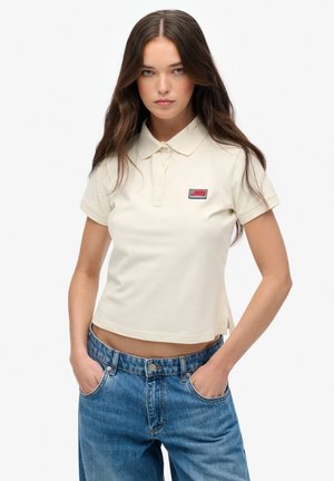 Lys beige kort polo-shirt med krave, korte ærmer og et lille broderet logo-patch på venstre bryst; parret med højttaljede blå jeans.