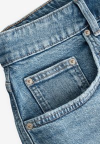 Gros plan sur un jean en denim bleu montrant la poche avant, la poche à pièces avec des rivets en métal, la passante de ceinture et les coutures détaillées.