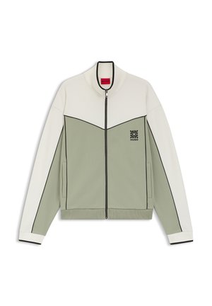 Jakke med lynlås, cremefarvet øvre del og ærmer, lysegrøn nederste del, sort kant, sidelommer og lille sort logo på brystet.