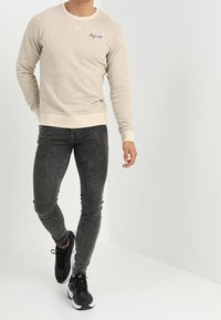 Sudadera de textura beige con puños y dobladillo acanalados, combinada con jeans ajustados grises y zapatillas deportivas negras. Detalle de logo bordado sencillo.