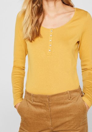 Vrouw die een mosterdgele henley top met lange mouwen en knopen draagt, gecombineerd met lichtbruine corduroy broek, handen in de zakken.